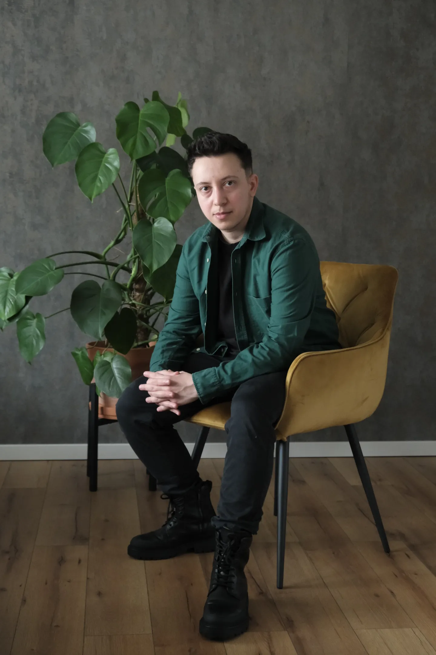 Young man in green shirt sitting on mustard chair beside a large potted plant on wood floor.