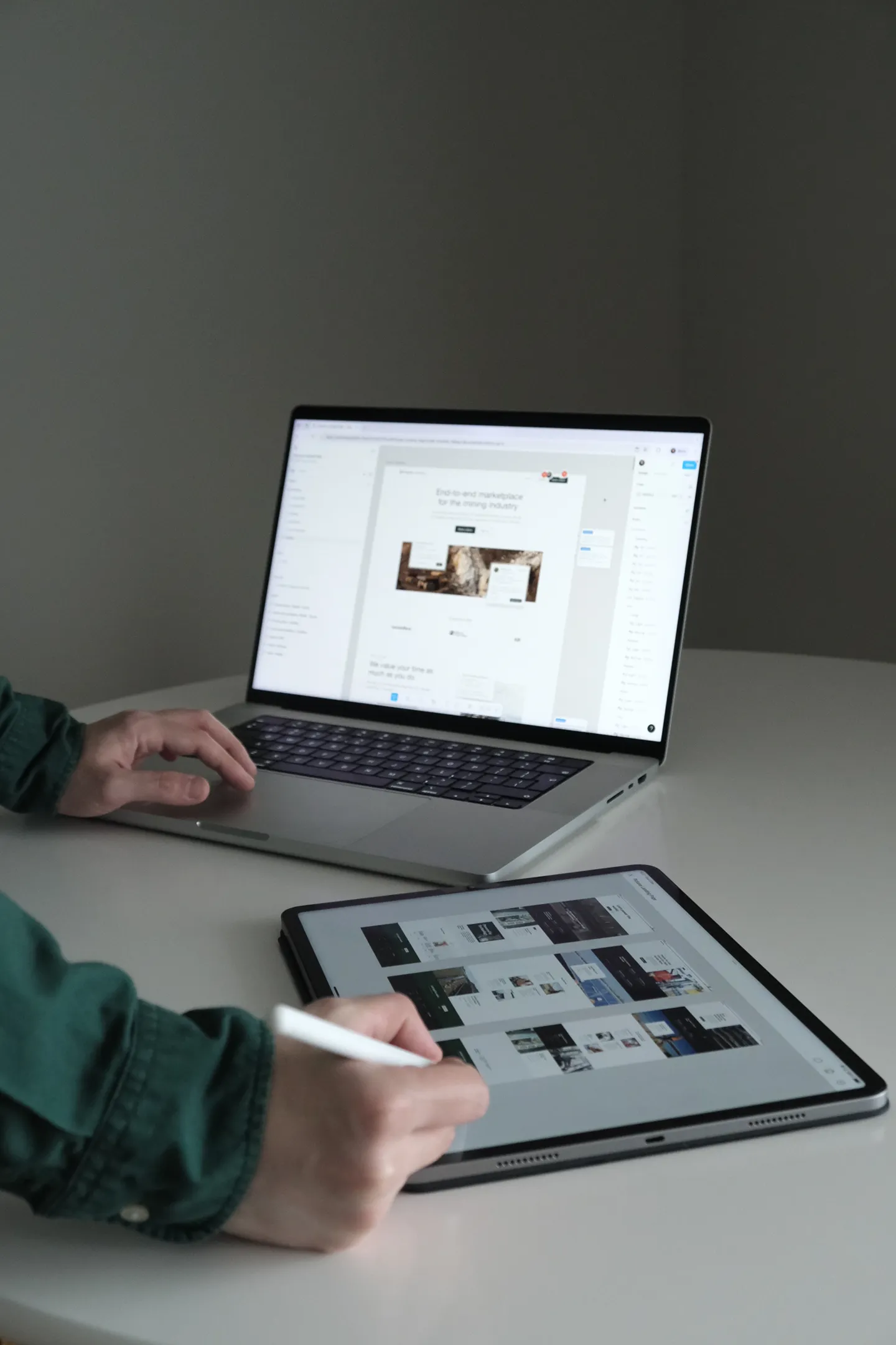 Person using stylus on tablet displaying design layouts, with laptop showing webpage design in background.