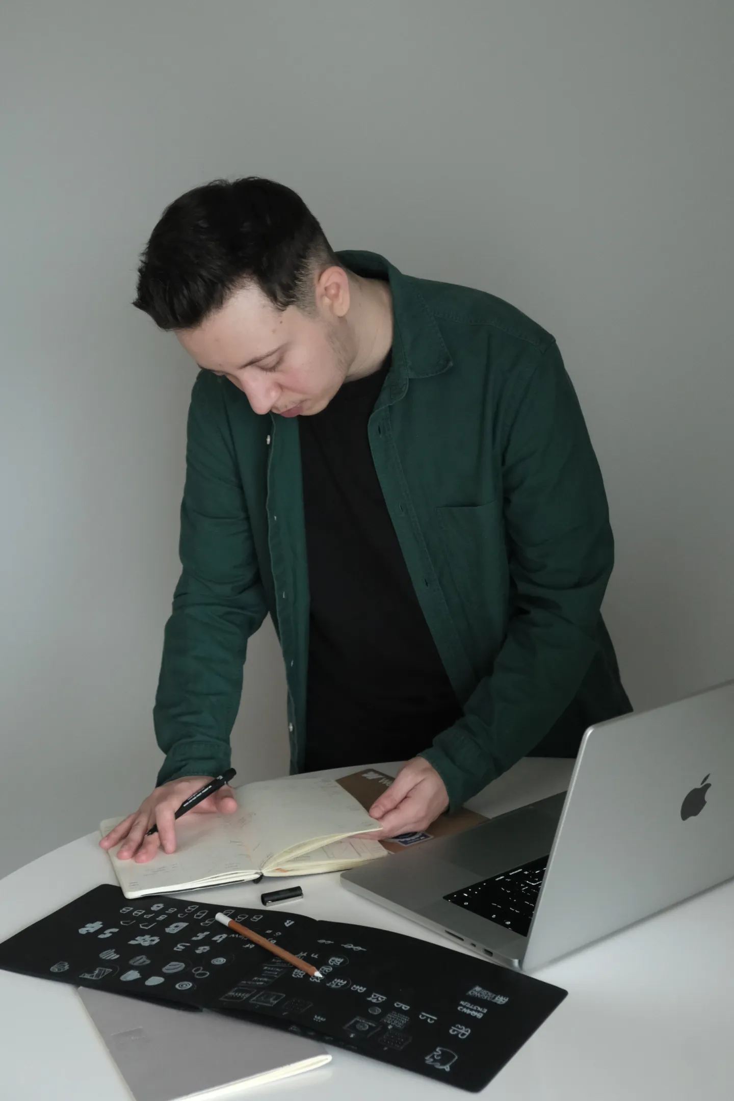 Young man in green shirt writing in a notebook at a desk with laptop and design sketches.